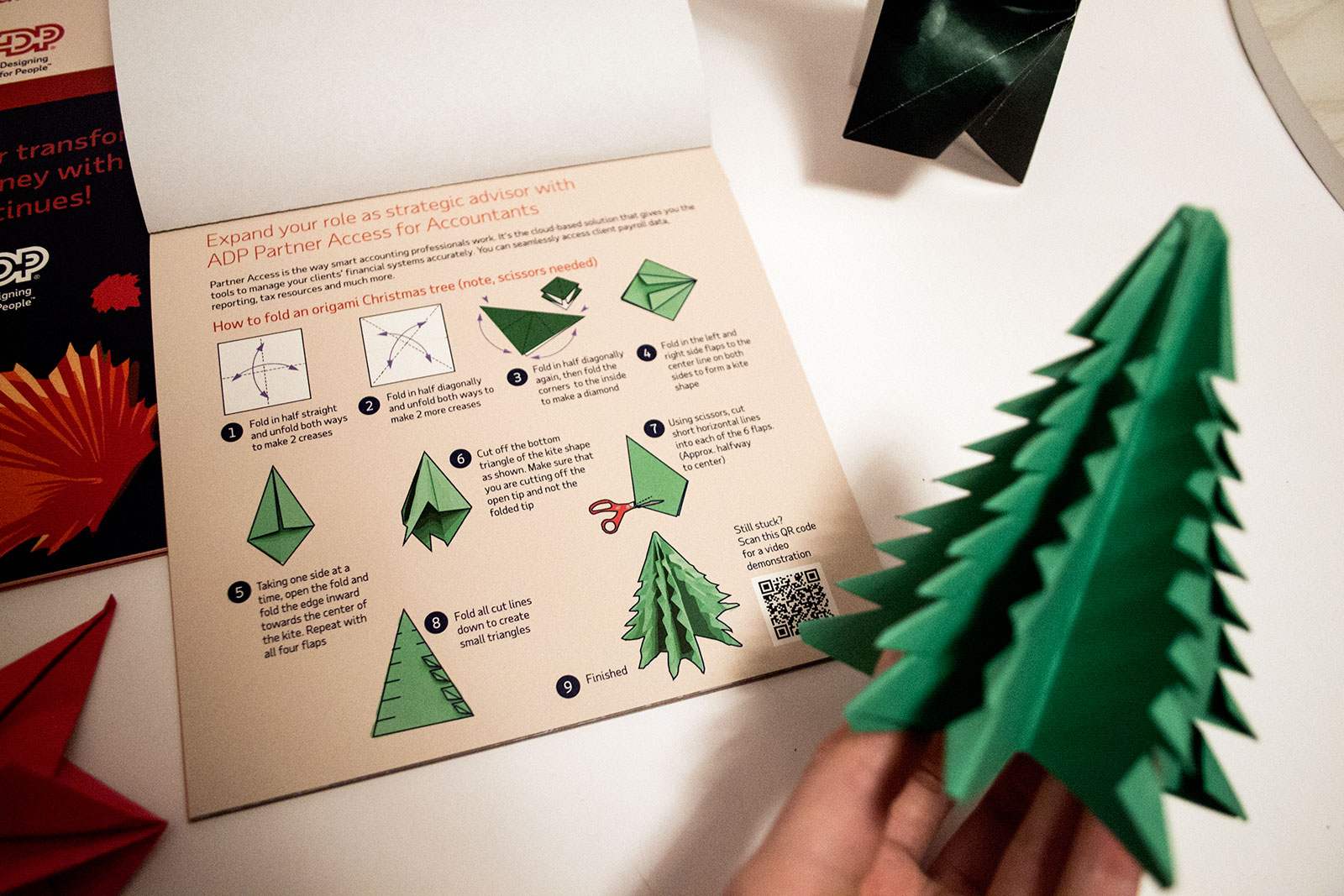 inside page instructions for folding an Origami christmas tree, with me holding a folded tree out of focus next to it