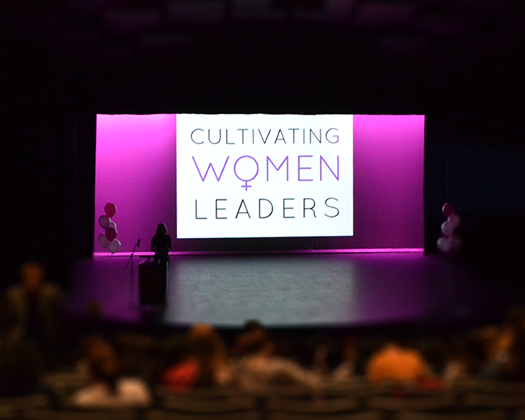 auditorium from the back row looking at the stage. The seats are packed and blurred out. The stage is illuminated with a pink light. There is a podium and a bunch of balloons to the left and on the projector screen is the Cultivating Women Leaders logo where the female symbol takes the place of the 'o' in women