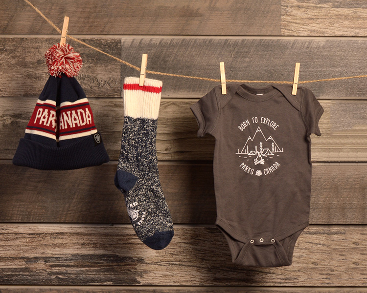 navy, white and red striped toque with PARKS CANADA embroidered in the stripe and a red and white pom-pom, navy sock sock and black onesie with Born to Explore Parks Canada illustration on the front, hanging on a clothesline