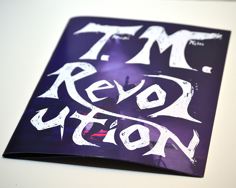 front cover of a book that reads with handdrawn letters 'TM Revolution' the background is in a dark indigo and you can make out a person standing behind the letters on a stage